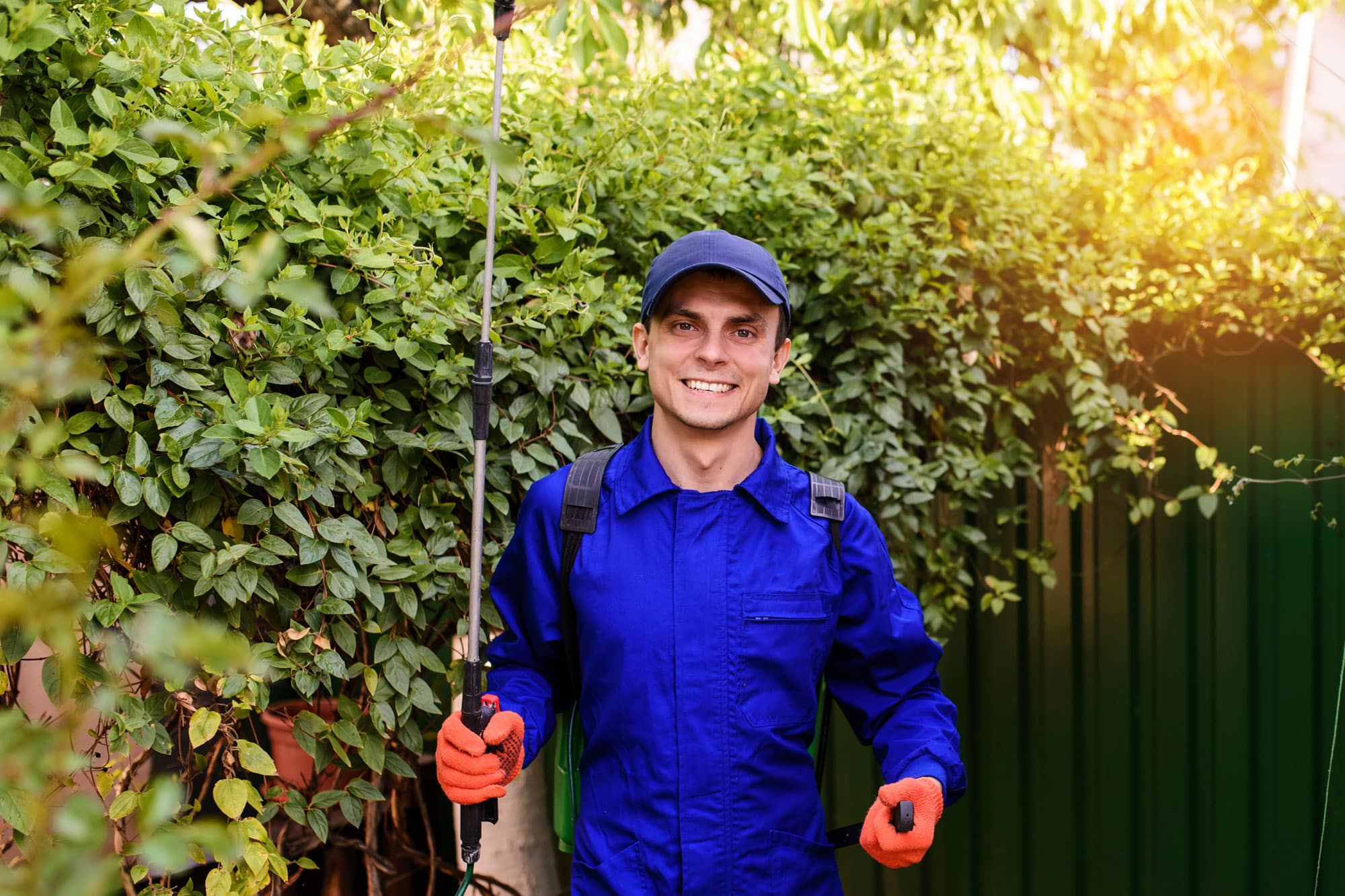 Pest Control expert in a blue shirt and gloves, pausing for a photo in the middle of spraying for insects on someone's property.